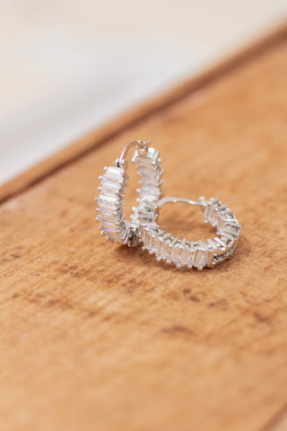 Pair of silver hoop earrings featuring baguette-cut clear crystals set in a full pavé design, displayed on a wooden surface. Glamorous and eye-catching earrings perfect for formal occasions or evening wear.