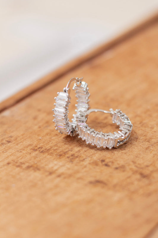 Pair of silver hoop earrings featuring baguette-cut clear crystals set in a full pavé design, displayed on a wooden surface. Glamorous and eye-catching earrings perfect for formal occasions or evening wear.