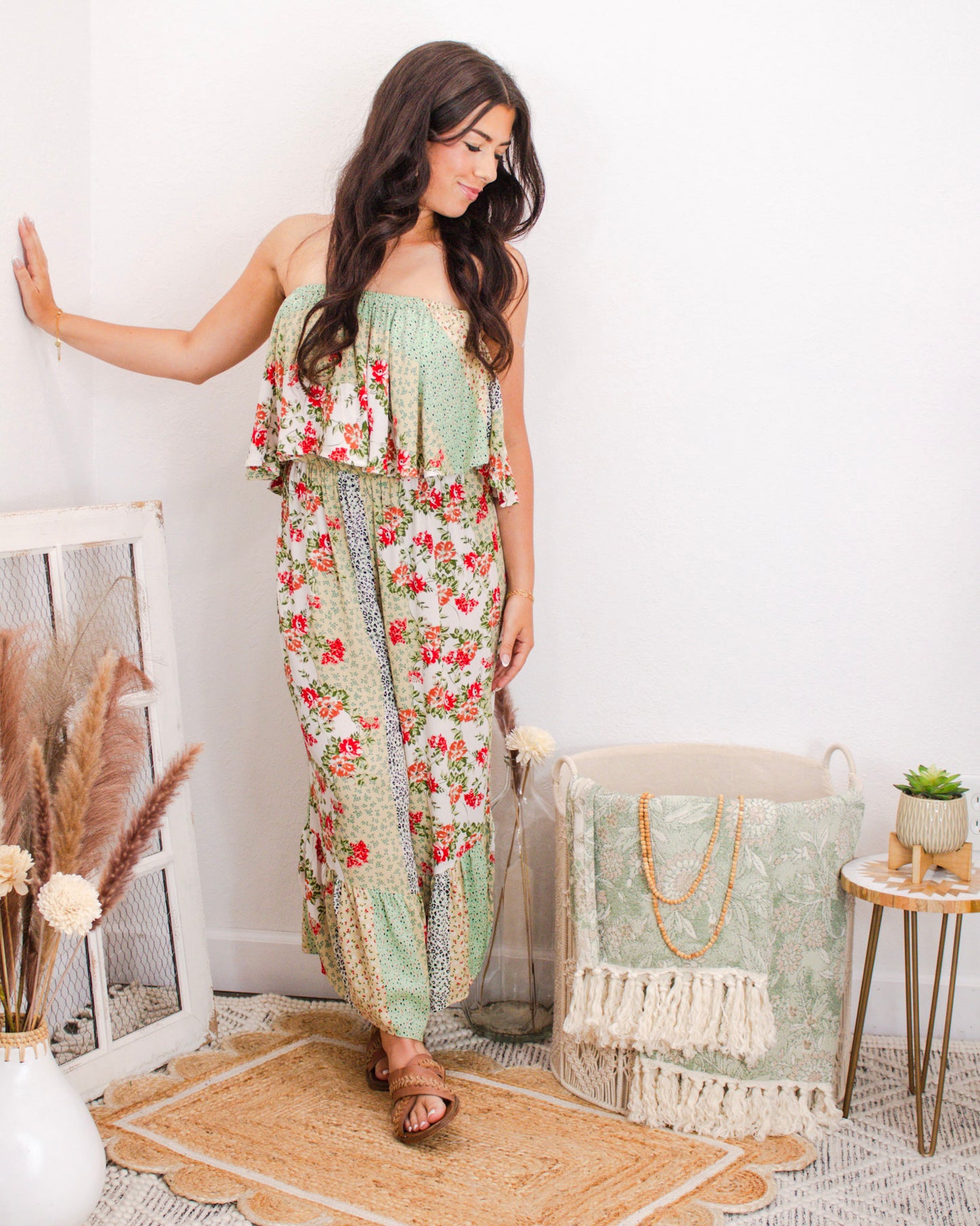 Woman wearing a strapless floral patchwork jumpsuit with wide legs and ruffled top, styled for summer in a boutique setting with neutral boho decor.