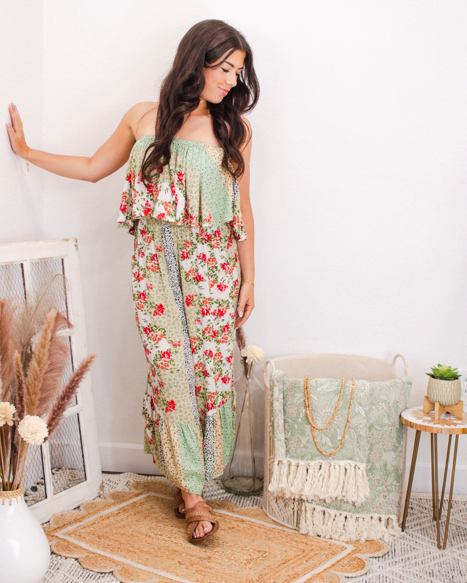 Woman wearing a strapless floral patchwork jumpsuit with wide legs and ruffled top, styled for summer in a boutique setting with neutral boho decor.