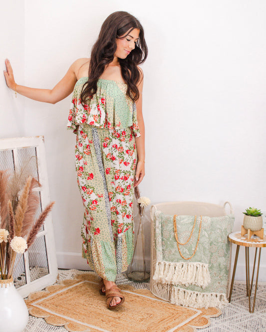 Woman wearing a strapless floral patchwork jumpsuit with wide legs and ruffled top, styled for summer in a boutique setting with neutral boho decor.