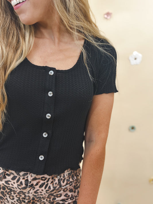 Woman wearing a black waffle knit short sleeve top with button front detail, styled with leopard print pants. Casual boutique fashion.