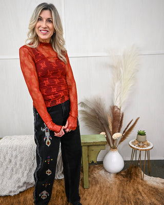 Model wearing a sheer long sleeve rust mesh top with floral embroidery and mock neckline, layered over a matching tank and paired with black denim embroidered jeans.