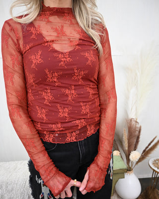 Close-up of a fitted long sleeve mock neck top in sheer rust mesh with embroidered floral lace overlay, styled over a matching cami and paired with black denim.