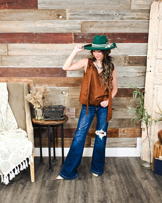 Model wearing a brown sleeveless tie-neck blouse with distressed wide-leg jeans, a green Western hat and statement necklace for a modern Western fall outfit.