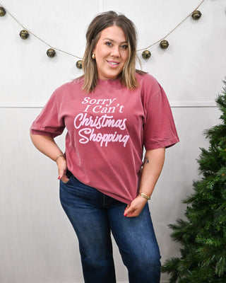 Woman wearing a maroon graphic T-shirt with the phrase ‘Sorry I Can’t Christmas Shopping,’ styled with dark-wash jeans and holiday décor.