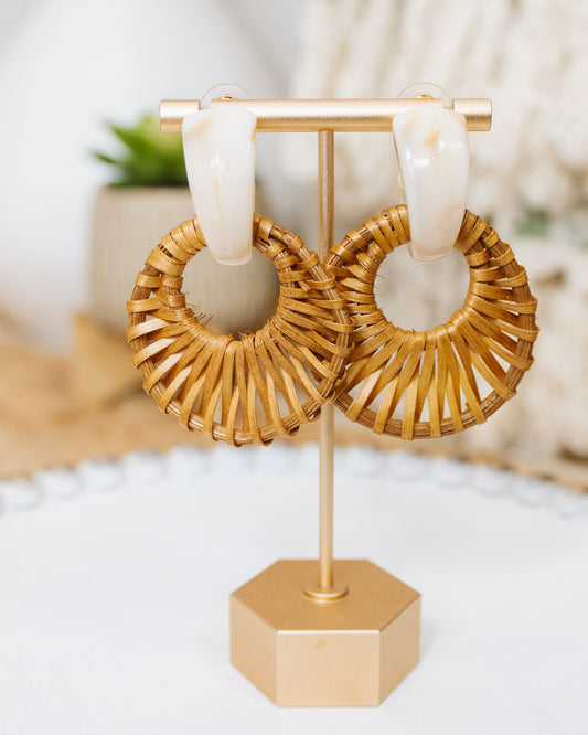 Pair of rattan woven hoop earrings with round natural wicker design and cream-colored acrylic posts, displayed on a gold earring stand with neutral boho decor.