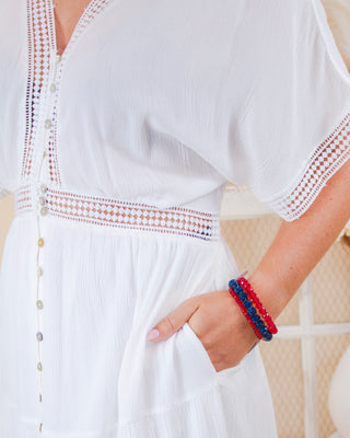 Close-up of woman wearing a white short sleeve dress with button-down front, eyelet lace trim, side pockets, and red and blue beaded bracelets
