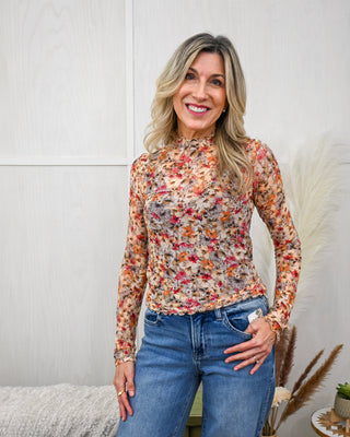 Model wearing a lightweight, sheer floral mock neck top with long sleeves in warm autumn tones, paired with mid-rise medium wash jeans for a casual chic look.