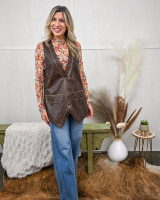 Model wearing a sleeveless faux leather brown vest with front pockets over a long sleeve floral mesh mock neck top, paired with relaxed-fit medium wash jeans.