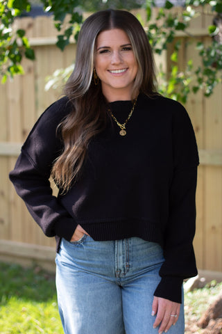 Smiling woman wearing a black cropped knit sweater with long sleeves, paired with light wash high-rise jeans and gold layered necklaces for a chic casual look.