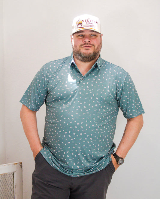 Man wearing a teal Raised By Coyotes polo with white Western-themed icons, paired with a white “Western Show” snapback hat and dark shorts.