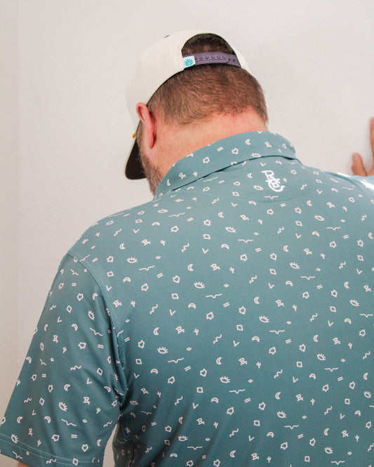 Back view of a man wearing a teal Sendero polo shirt with white desert-themed symbols and a branded collar, paired with a white snapback hat.