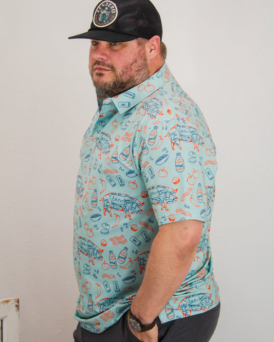 Man wearing a teal short sleeve Sendero shirt with BBQ-inspired graphics and a black mesh trucker hat featuring a round Sendero Provisions Co. embroidered patch.