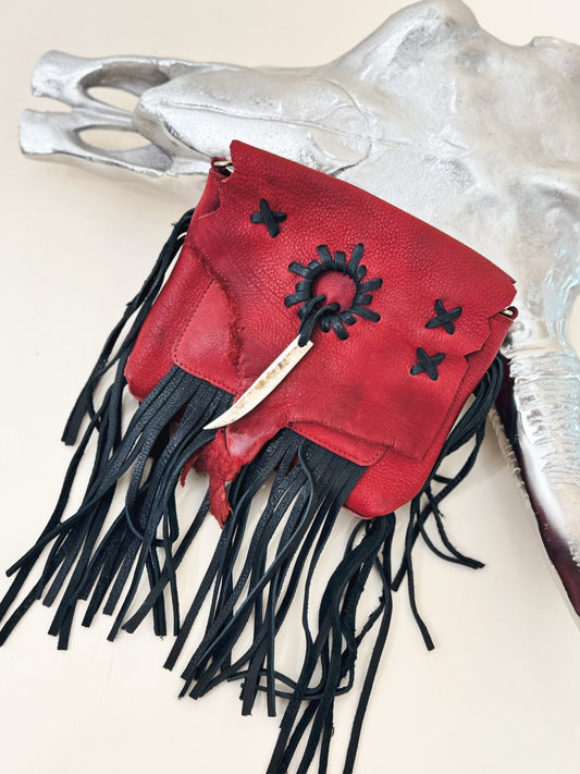 Red leather crossbody pouch with black fringe, hand-stitched details, and antler accent, styled on a silver skull for a bold Western-inspired statement.