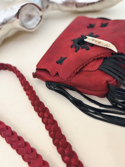Close-up of a red leather crossbody pouch with braided strap, black fringe, and antler detail—Western-inspired handcrafted accessory with rustic charm.