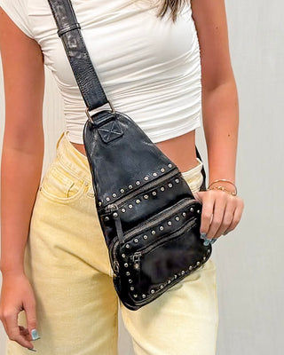 Black studded leather sling backpack with multiple zippered compartments and adjustable crossbody strap, styled with yellow denim shorts and a fitted cream top for an edgy boutique look.