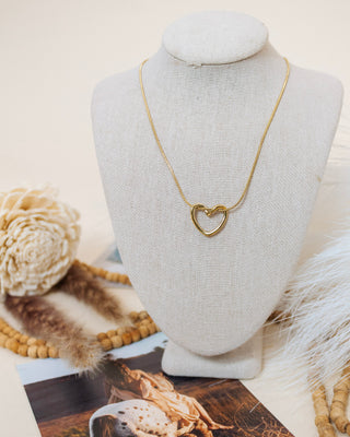 Gold open-heart pendant necklace on a fine snake chain, displayed on a neutral linen bust with wooden bead and pampas grass accents in the background