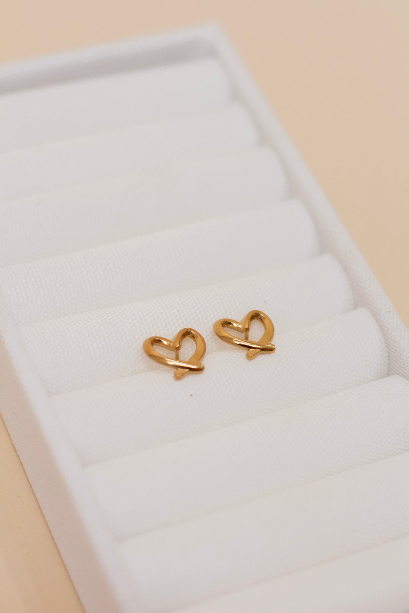 Pair of small gold heart-shaped stud earrings with an open design and subtle curve, displayed on a white fabric surface. Dainty and minimalist jewelry perfect for everyday wear or gifting.