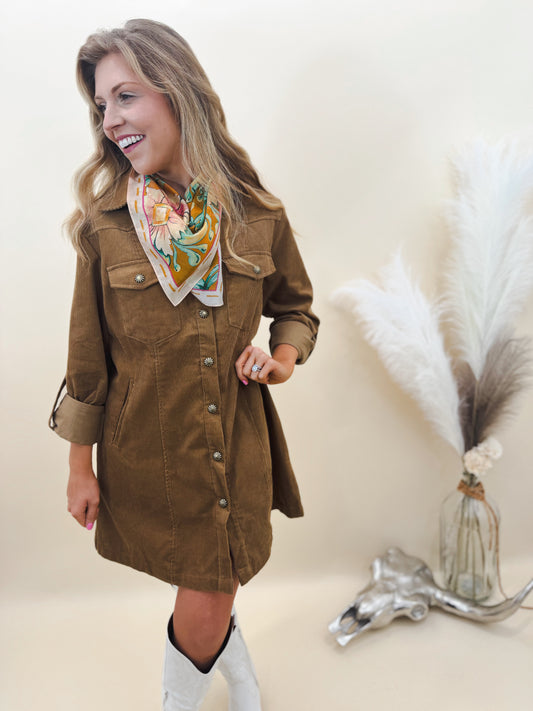 Woman wearing a brown corduroy shirt dress with floral bandana scarf and white cowboy boots, styled for a western-chic look with rolled sleeves and silver snaps.