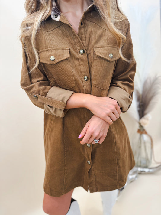 Close-up of woman wearing a brown corduroy shirt dress with rolled sleeves, front pockets, and silver concho-style snap buttons, paired with white western boots.