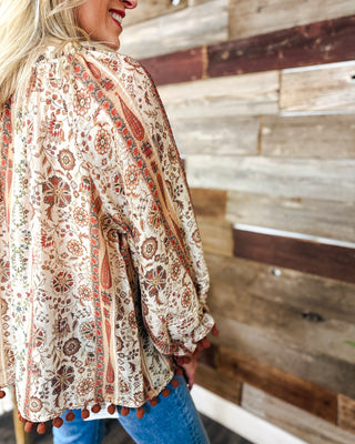 Close-up of women’s cream boho floral blouse with rust paisley print, relaxed balloon sleeves and pom-pom trim in warm earthy fall tones.