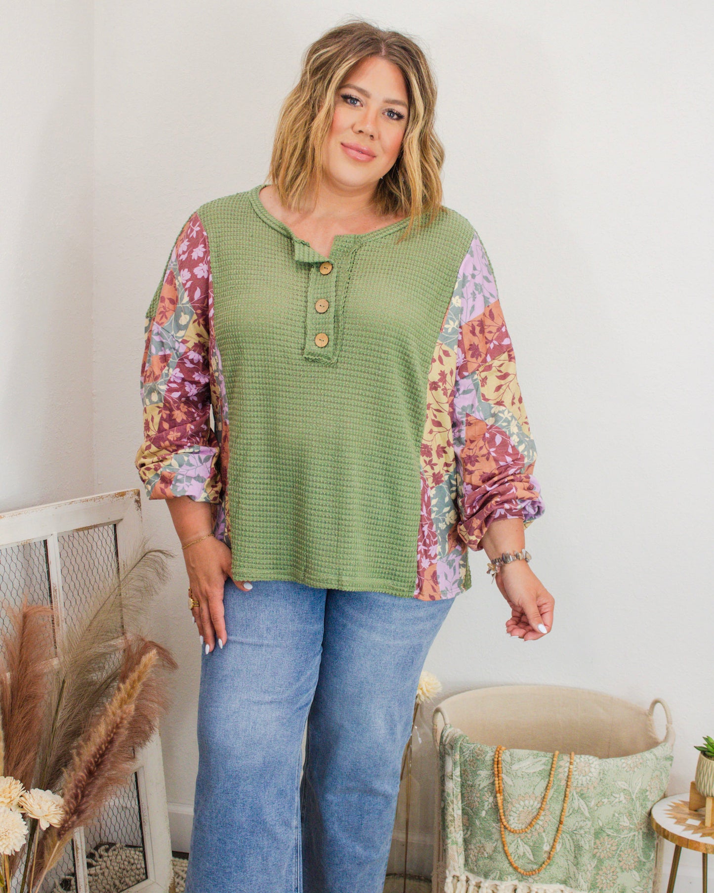 Plus-size woman modeling a green waffle knit henley top with colorful floral patchwork sleeves, paired with light-wash denim jeans in a boutique setting.