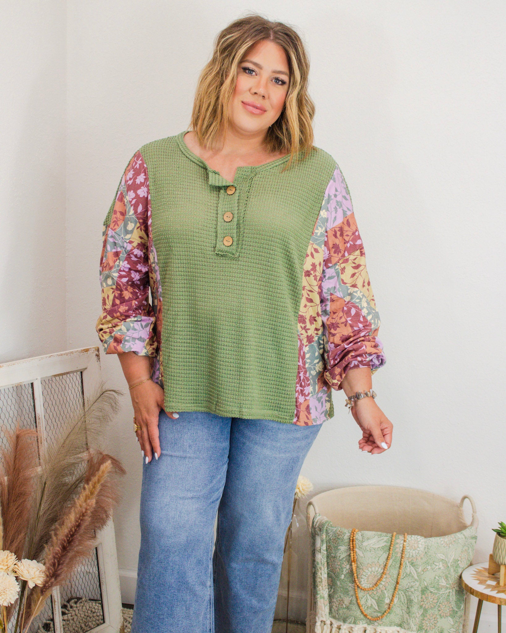 Plus-size woman modeling a green waffle knit henley top with colorful floral patchwork sleeves, paired with light-wash denim jeans in a boutique setting.