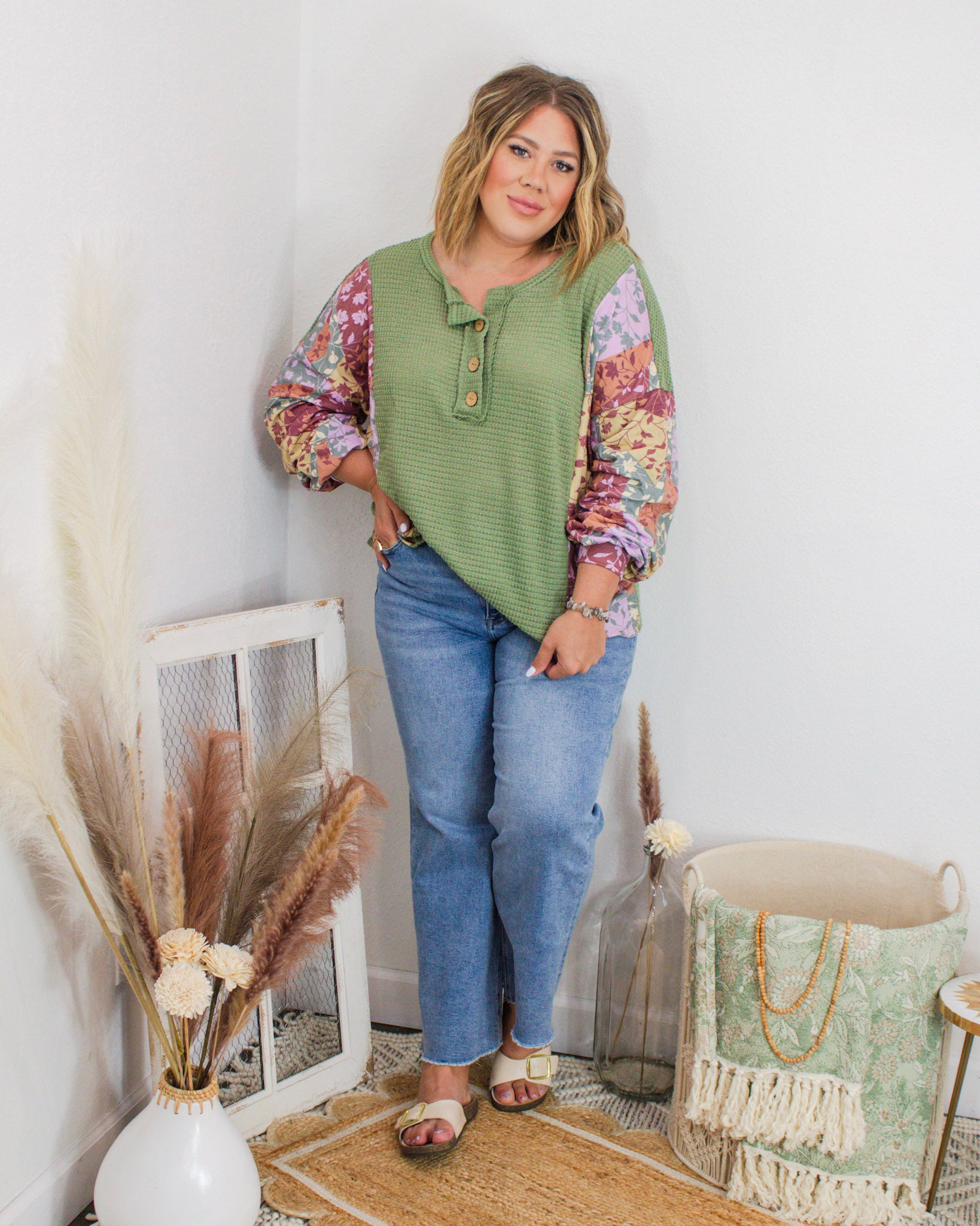Woman wearing a green waffle knit henley top with floral patchwork sleeves, styled with denim jeans and sandals in a boho boutique setting.