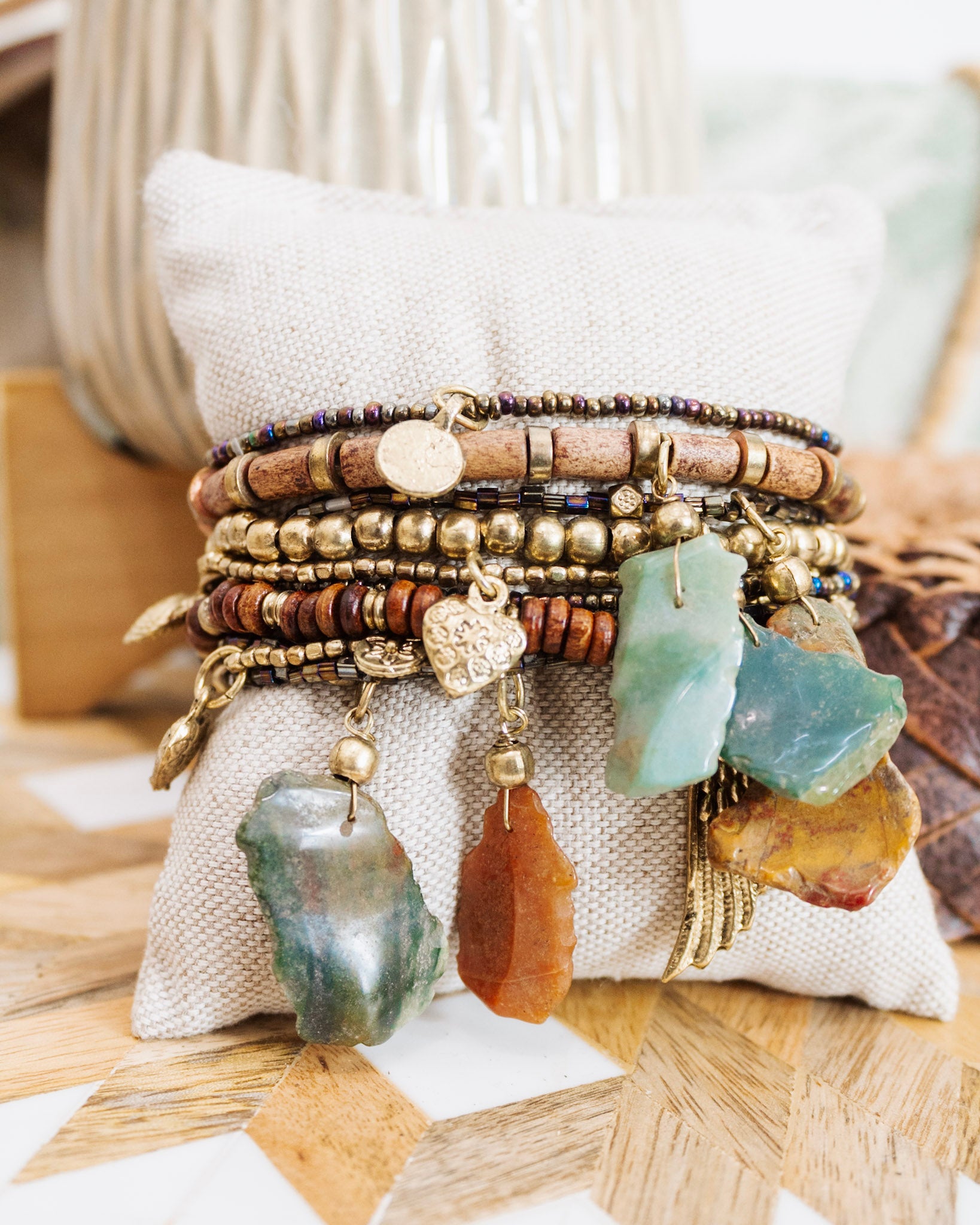 Stack of boho gemstone charm bracelets with mixed beads, wood accents, and gold detailing displayed on a neutral cushion.