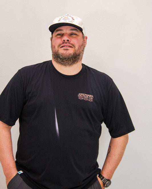 Man wearing a black Sendero t-shirt and a white trucker hat with graphic text, standing casually against a plain background with hands in pockets.
