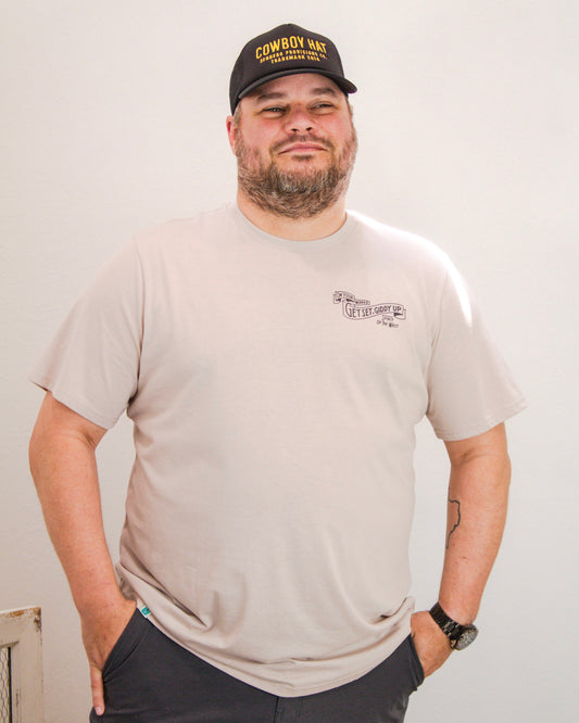 Man wearing a light beige Sendero Provisions Co. t-shirt with “Get Set, Giddy Up” graphic on the chest, paired with a black “Cowboy Hat” trucker hat.