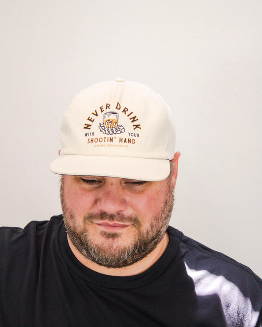 Man wearing cream Sendero Provisions Co. hat with “Never Drink with Your Shootin’ Hand” text and skeleton hand holding a drink graphic on the front.