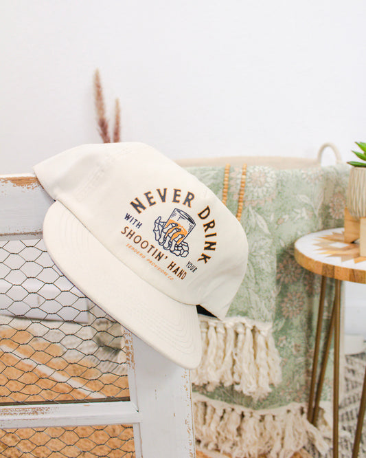 Cream-colored Sendero Provisions cap with “Never Drink with Your Shootin’ Hand” text and skeleton hand graphic, displayed on rustic white chicken wire frame.