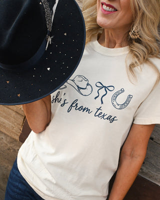 Cream western graphic tee with “She’s From Texas” design styled with dark wash flare jeans and a black rancher hat for trendy boutique western fashion.
