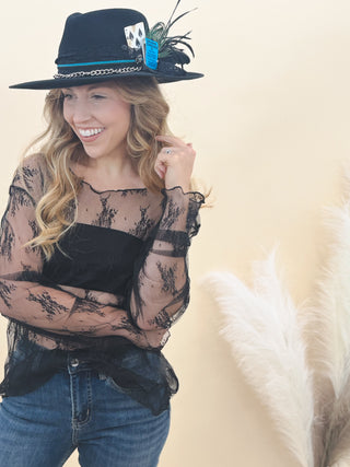 Woman wearing a sheer black lace top, dark denim jeans, and a stylish black wide-brim hat adorned with feathers, cards, and a blue tag, standing against a neutral background.