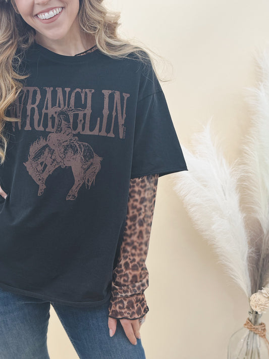 Woman wearing a black “WRANGLIN” graphic tee layered over leopard print sleeves, paired with denim jeans for a bold, western-inspired look.
