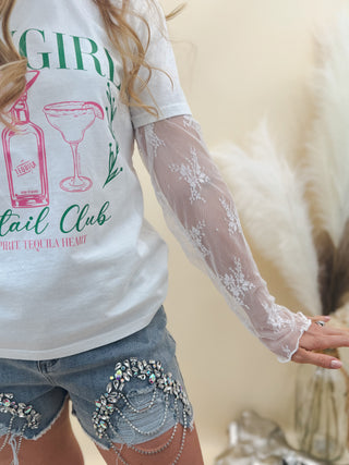 White "Tequila Girl Cocktail Club" graphic tee layered over sheer lace sleeves, styled with high-rise denim shorts adorned with rhinestones and chain fringe.