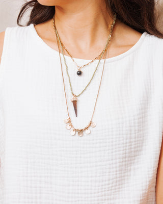 Close-up of a woman wearing a layered necklace set with gold chain and black bead, green beaded strand with spike pendant, and gold disc charm necklace over a white textured top.