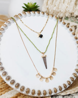 Flat lay of three layered necklaces on a round white beaded-edge stand featuring a gold chain with black charm, green beaded strand with spike pendant, and gold disc charms.