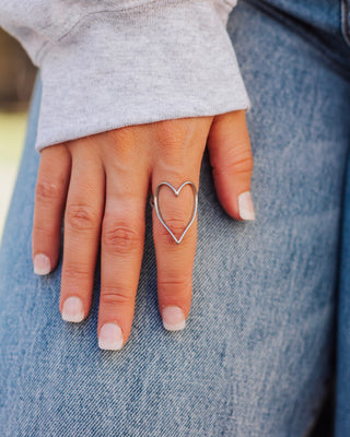 Dainty silver heart ring with an open wire design, styled with a gray sleeve and blue denim.