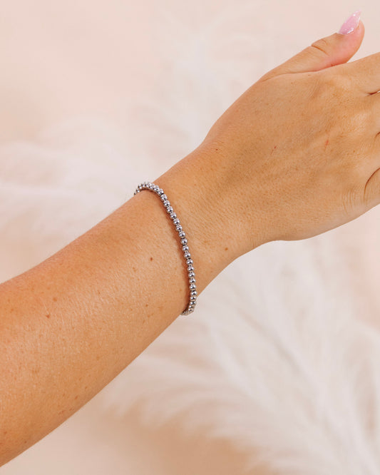 Close-up of a woman's wrist wearing a silver tennis bracelet with shimmering stones, styled against a soft blush background and manicured pink nails.
