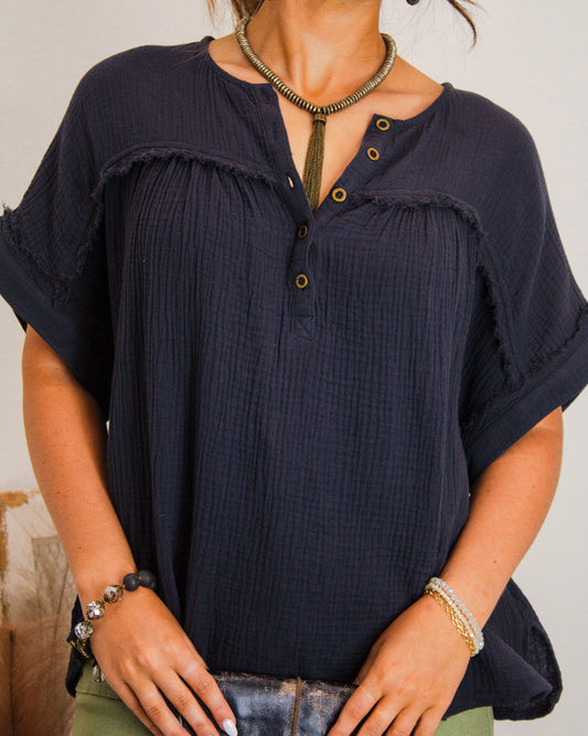 Close-up of a woman wearing a black textured button henley top with raw edge detailing, accessorized with a tassel necklace and layered bracelets.