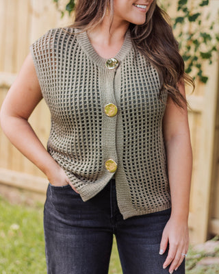 Woman wearing a sleeveless olive green knit cardigan with large gold buttons, styled with dark wash jeans for a chic and effortless fall outfit.
