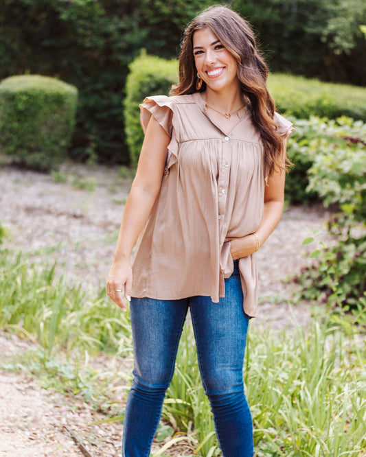 Woman wearing a taupe button-front blouse with flutter sleeves, styled with skinny blue jeans. Lightweight and flowy neutral top for casual or dressy outfits.