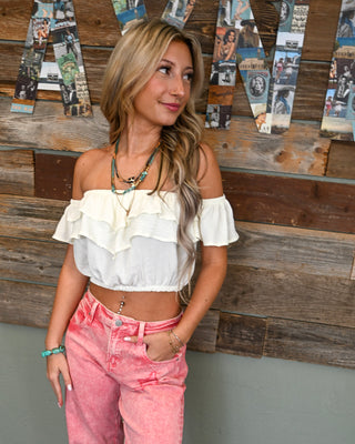 Women’s ivory off-the-shoulder ruffle crop top with layered flutter sleeves, styled with pink mineral wash jeans for a playful boho boutique look.