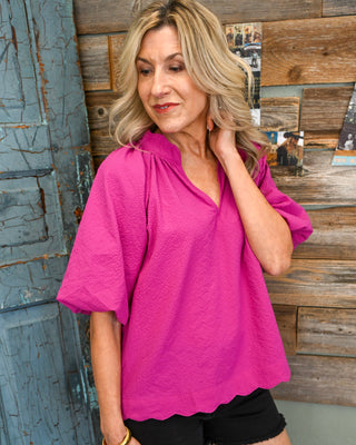 Women’s hot pink textured blouse with split neckline, puff sleeves, and scalloped hem, styled with black frayed shorts for a bright boho boutique look.