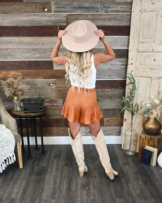 Rust orange pleated mini skirt with ruffle hem, styled with a white tank, cowboy boots and wide brim hat for a trendy western fall outfit.
