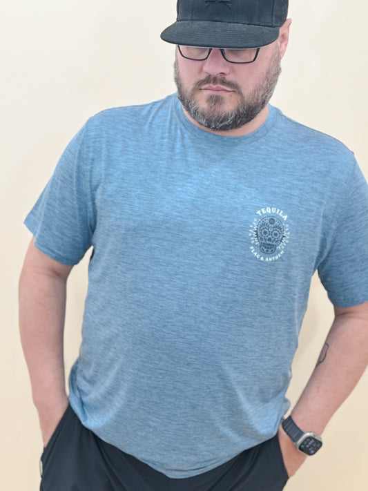 Man wearing a heather blue “Tequila Made Me Do It” graphic tee with a sugar skull design, black athletic shorts, black hat, and smartwatch.