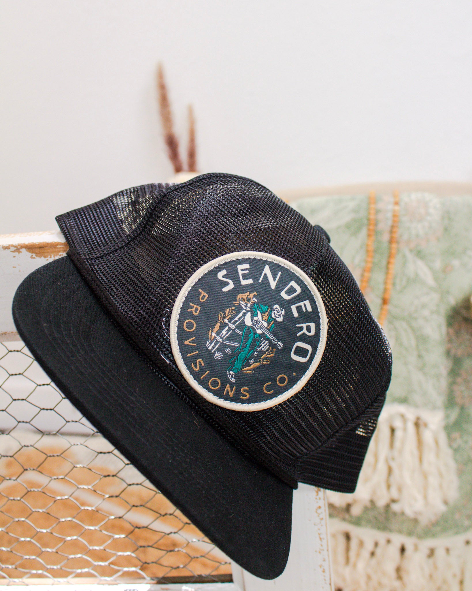 Black mesh trucker hat from Sendero Provisions Co. with a round patch featuring a cowboy illustration, displayed on a rustic backdrop with neutral decor.